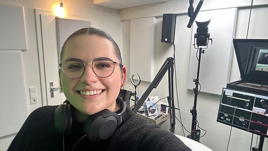 Blick in das Film- und Tonstudio Das Bild zeigt eine Frau in einem Studio. Um den Hals hat sie einen Kopfhörer. Im Hintergrund sieht man Equipment wie Mikrophon, Lichter, Computer, Kamera.