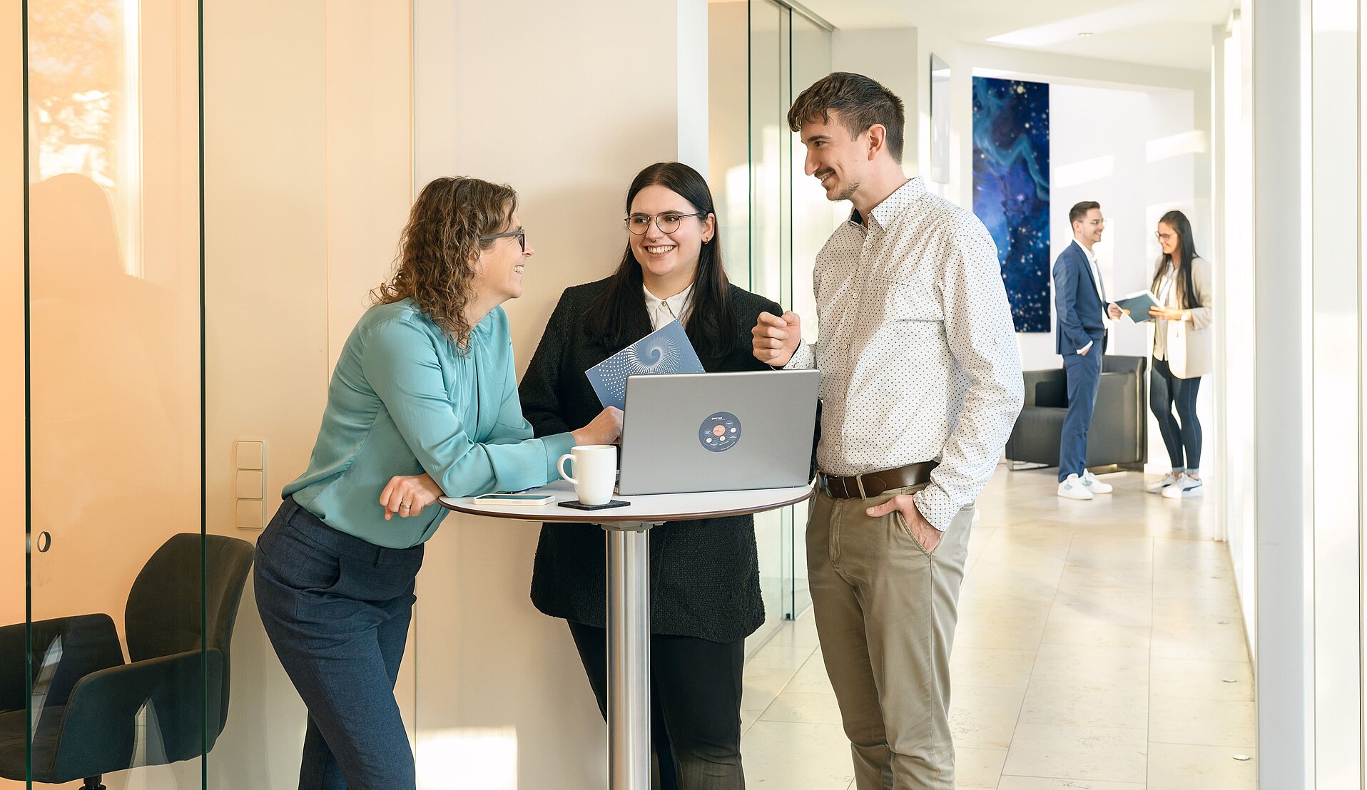 Teamwork am Stehtisch Drei Kollegen – zwei Frauen und ein Mann – stehen an einem Stehtisch im modernen Büroflur von Corivus und diskutieren angeregt über Inhalte auf einem geöffneten Laptop. Im Hintergrund sind zwei weitere Kollegen schemenhaft im Gespräch zu erkennen. Die Umgebung ist offen und hell gestaltet, mit Glaswänden und modernen Einrichtungselementen.