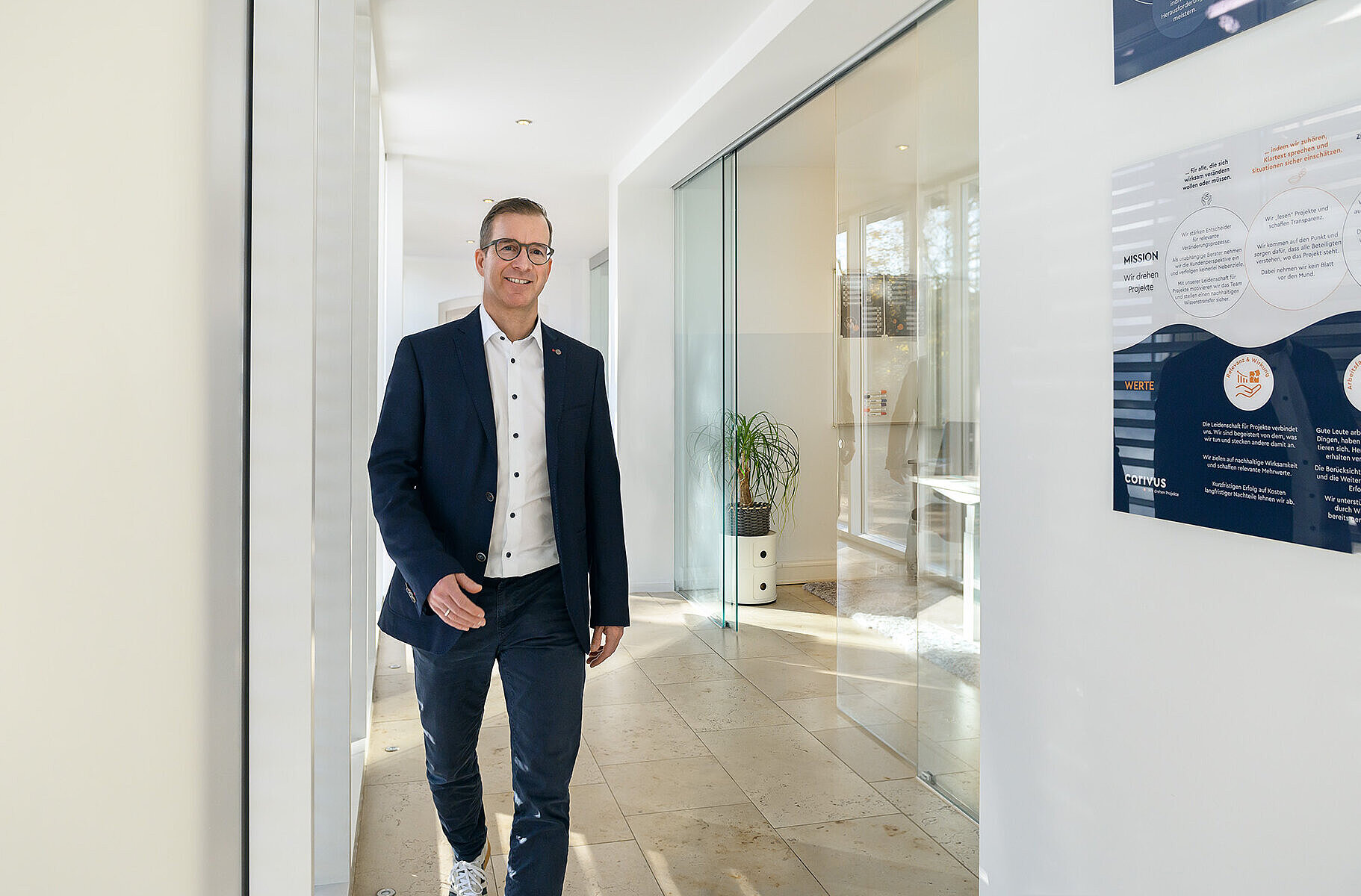 Christian Dürk, Vorstand der Corivus AG – Projektmanagementberatung Das Bild zeigt Christian Dürk, Vorstand der Corivus AG für Projektmanagementberatung, im modernen Hauptsitz in Neustadt an der Weinstraße. Er geht durch einen lichtdurchfluteten Flur mit Glaswänden, auf denen die Mission und Werte der Corivus AG dargestellt sind. Die Szene vermittelt Professionalität, Transparenz und die beratungsorientierte Unternehmenskultur der auf Projektmanagement spezialisierten Unternehmensberatung.