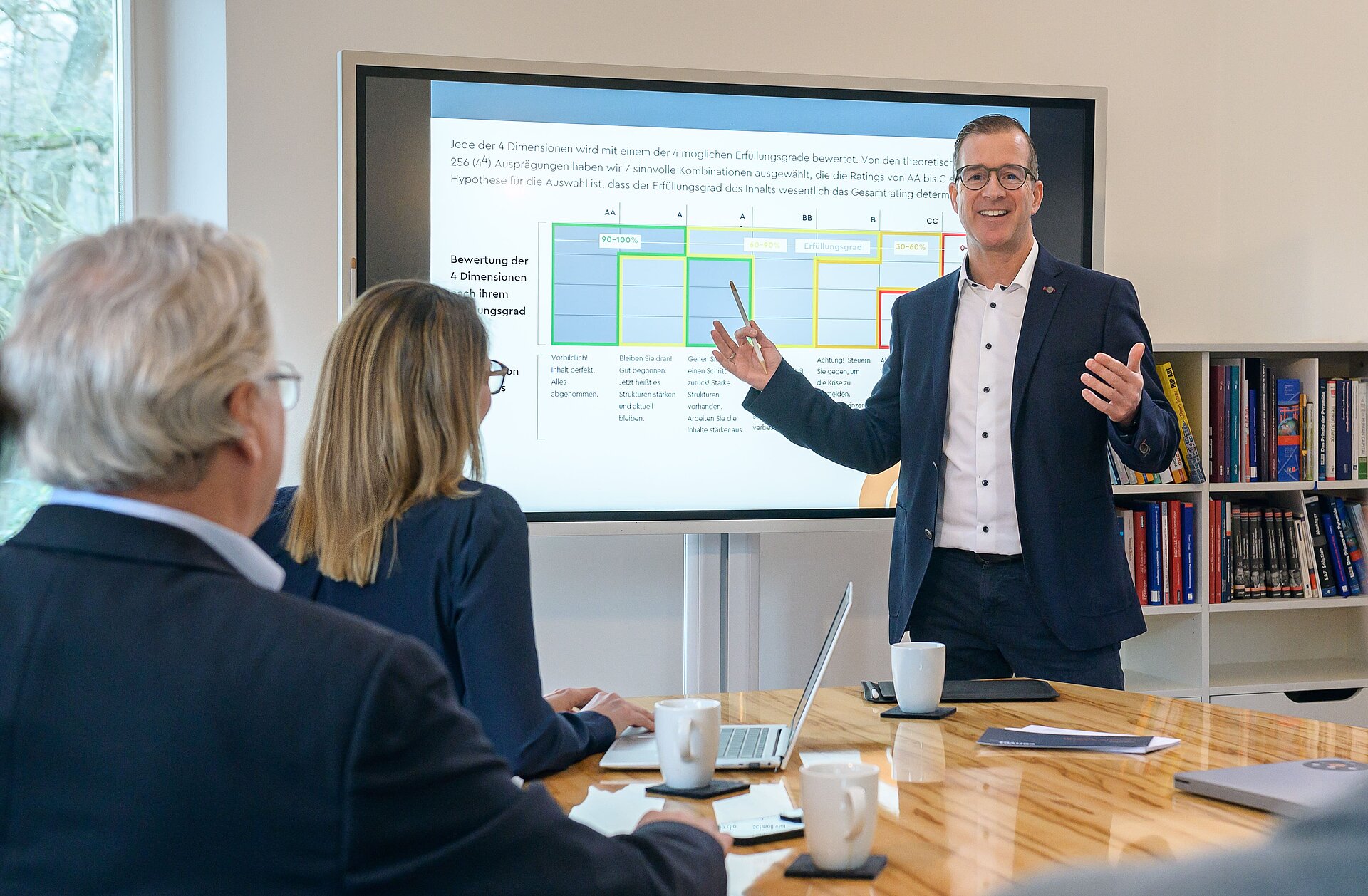 Dynamischer Vortrag in professioneller Besprechung Christian Dürk hält eine Präsentation im modernen Konferenzraum von Corivus. Mehrere Meetingteilnehmer sitzen an einem großen Konferenztisch aus Holz, hören aufmerksam zu und nehmen aktiv an der Diskussion teil. Notizbücher und Getränke auf dem Tisch zeigen eine produktive Arbeitsatmosphäre.