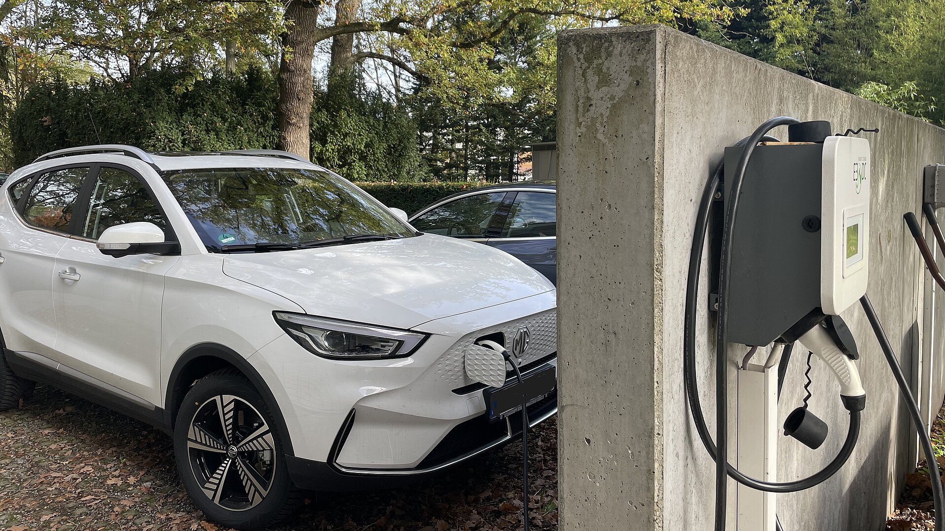 Laden während der Arbeitszeit Das Bild zeigt ein weißes Elektro-Fahrzeug. Es steht auf einem Parkplatz. An der Wand ist eine Wallbox montiert. Das Auto lädt gerade Strom. Der Stecker ist eingesteckt.