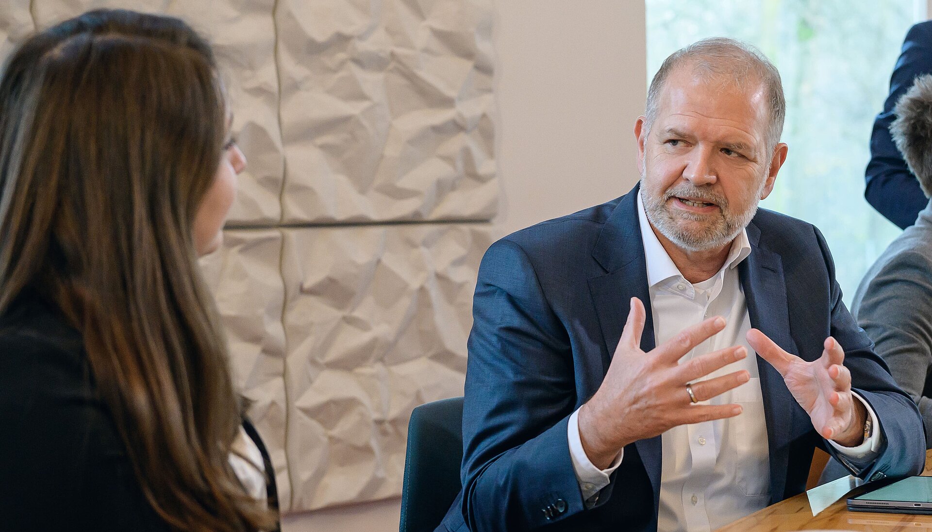 Erfahrungsbasierter Austausch in der Business-Diskussion Ein erfahrener Geschäftsmann im dunkelblauen Anzug spricht leidenschaftlich mit einer Kollegin, während er mit ausdrucksstarker Gestik seine Argumente unterstreicht. Der Besprechungsraum mit modernen Akustikpaneelen im Hintergrund schafft eine professionelle Atmosphäre.